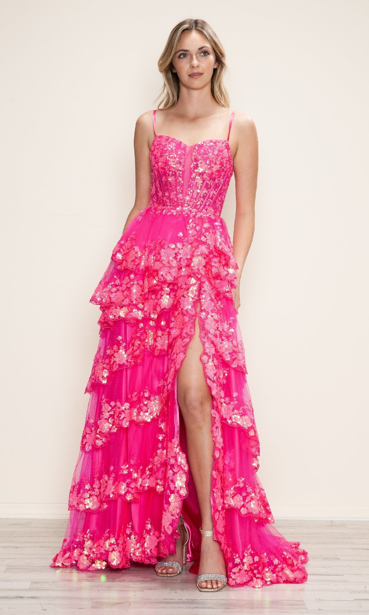 A woman poses against a plain background in the Long Prom Dress: Poly USA 9606— a bright pink, tiered A-line gown with floral appliqué, spaghetti straps, and a thigh-high slit—paired with silver open-toe heels.